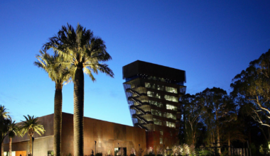 A modern building with an angular glass tower and palm trees is illuminated against a deep blue twilight sky.