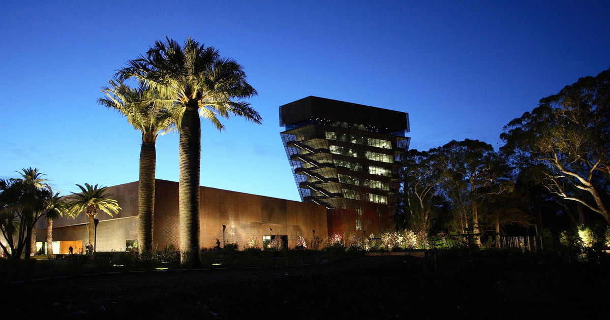 A modern building with an angular glass tower and palm trees is illuminated against a deep blue twilight sky.