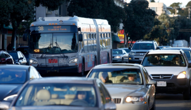 A city bus labeled "38 Geary Transbay Terminal" and several cars are in heavy traffic on a tree-lined urban street during daylight.