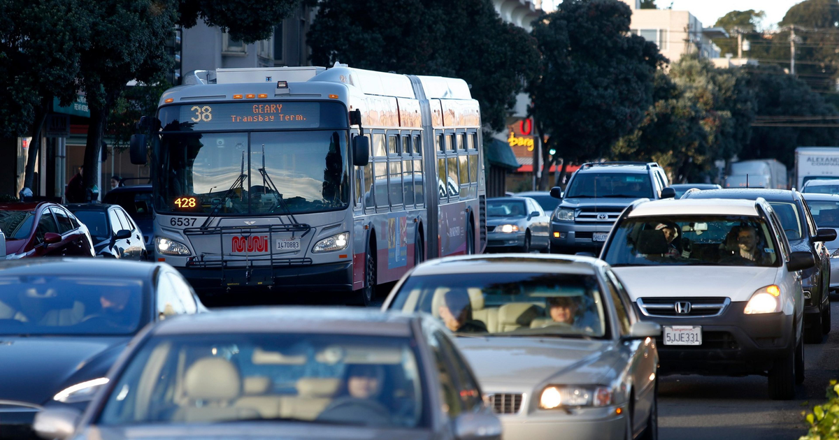 A city bus labeled "38 Geary Transbay Terminal" and several cars are in heavy traffic on a tree-lined urban street during daylight.