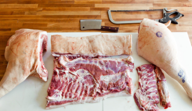 A man with a tattooed arm sharpens a knife while standing next to a large cut of raw pork on a metal table.