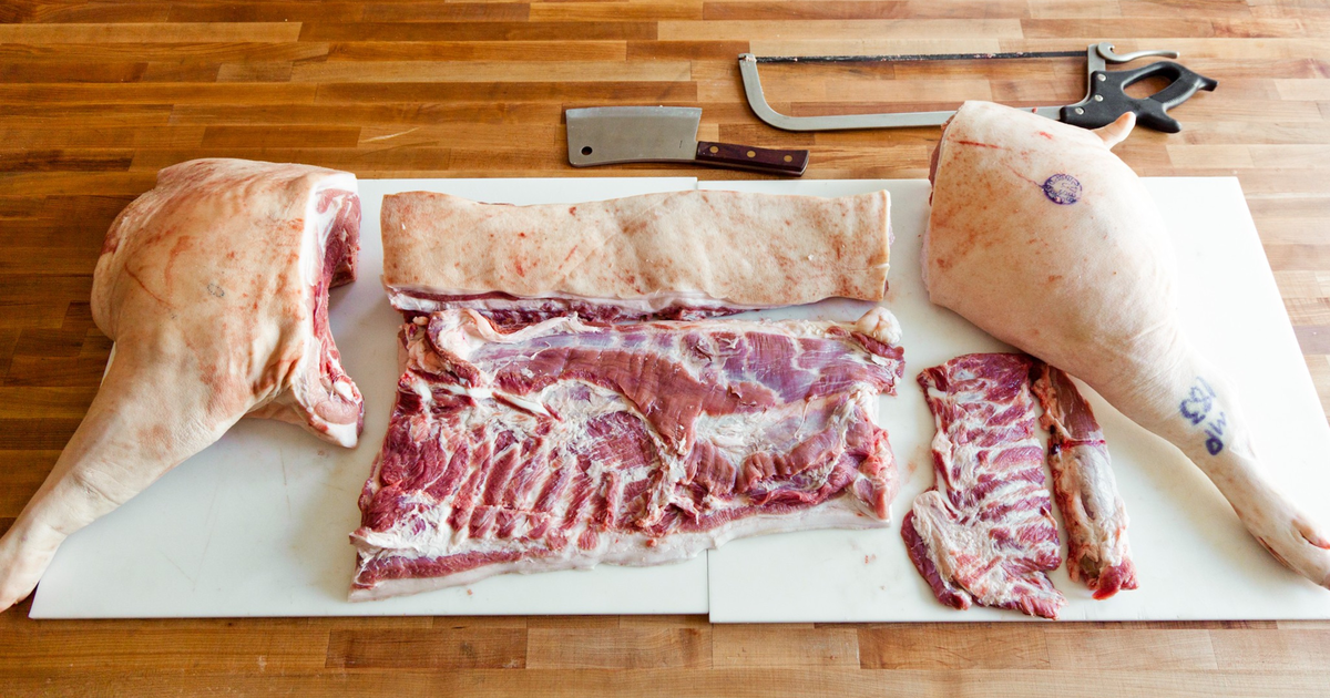 A man with a tattooed arm sharpens a knife while standing next to a large cut of raw pork on a metal table.