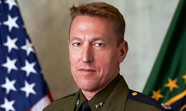 A headshot of a person in uniform standing in front of two flags. The one on the left is the U.S. flag.