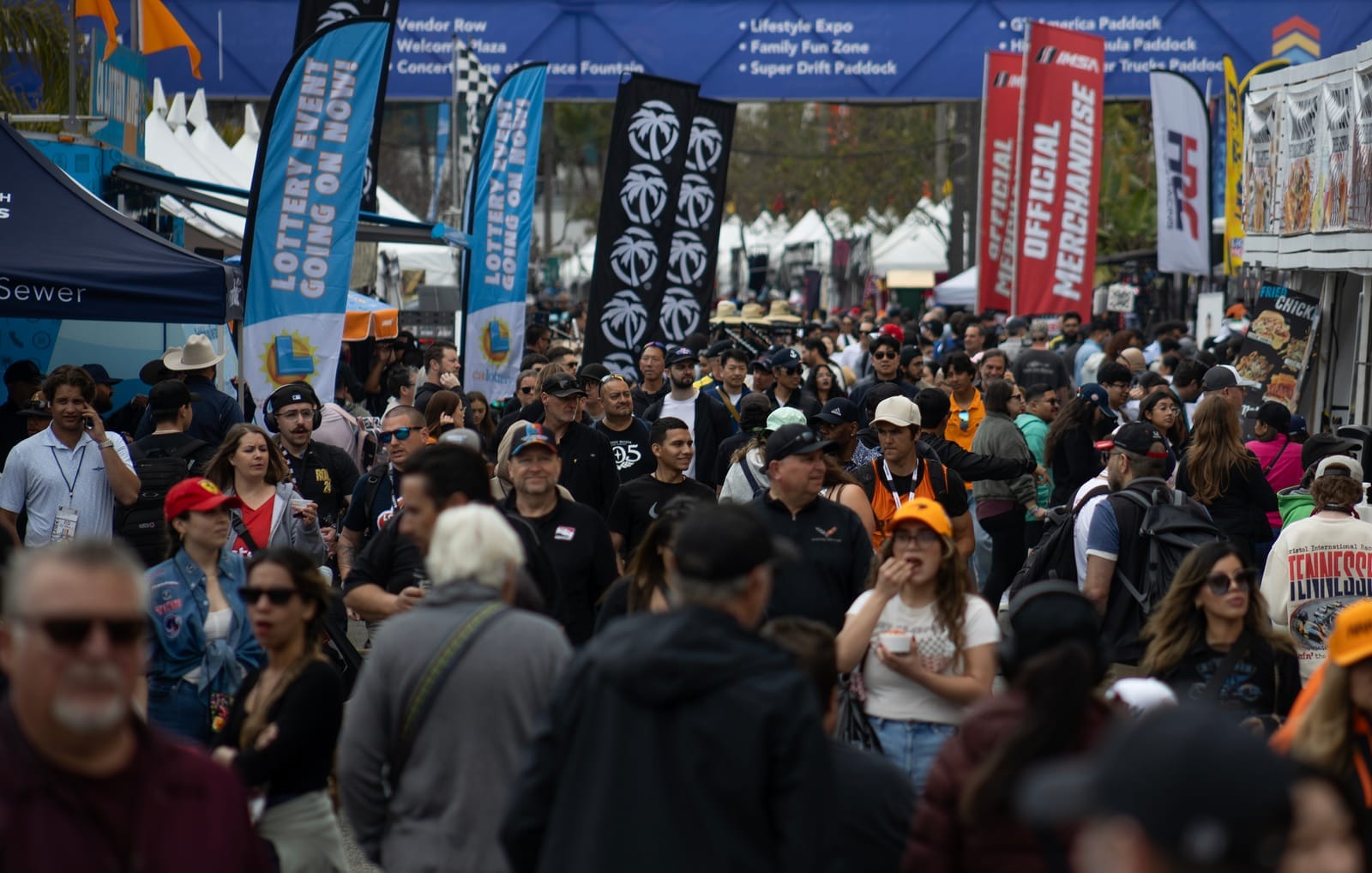 A large crowd of people walk shoulder to shoulder outside beneath colorful advertising banners.