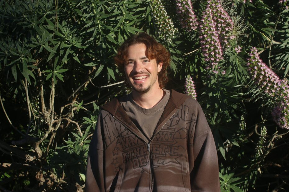 A man with medium-length brown hair and a beard smiles while standing in front of green foliage and purple flowers, wearing a brown hoodie.