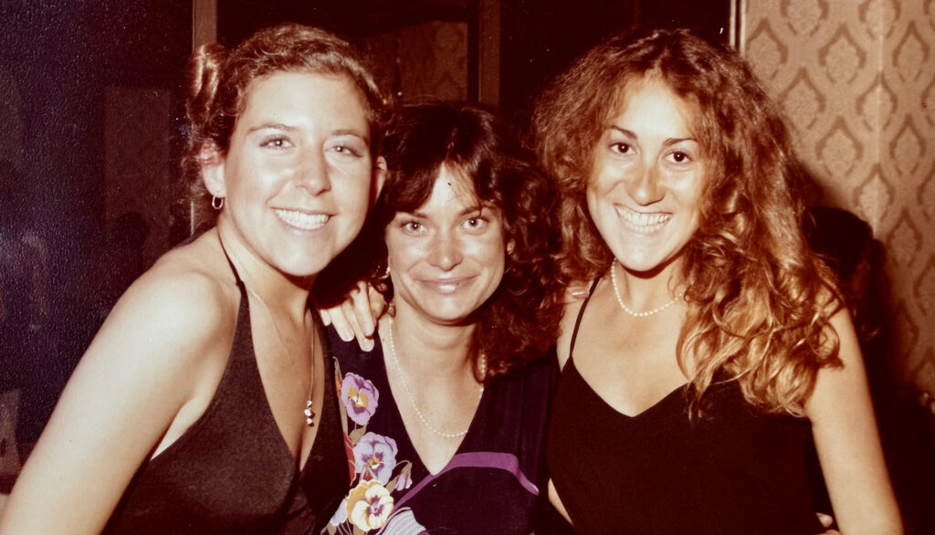 A vintage, warm-toned photo of three smiling young women, including Cathy Sandrich Gelfond, posing closely together. They are dressed in evening wear, capturing a candid and joyful moment at a social gathering.
