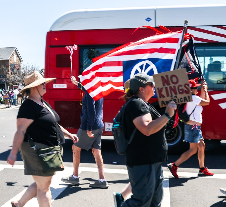 People march on a sunny street carrying flags and a sign reading "No Kings in America." A red bus is visible in the background, adding vibrant color.