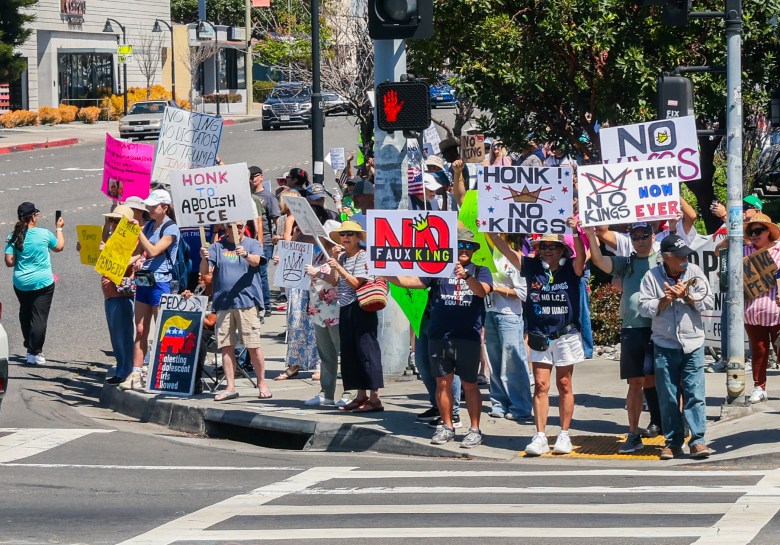 Protesters at a street corner hold various anti-authority signs, including "No Kings" and "Abolish ICE," conveying a message of resistance.