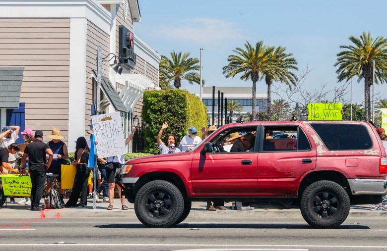 People protest on a sunny day by a building with palm trees. They hold signs against war, including "No ICE, No War," and a red SUV passes by.