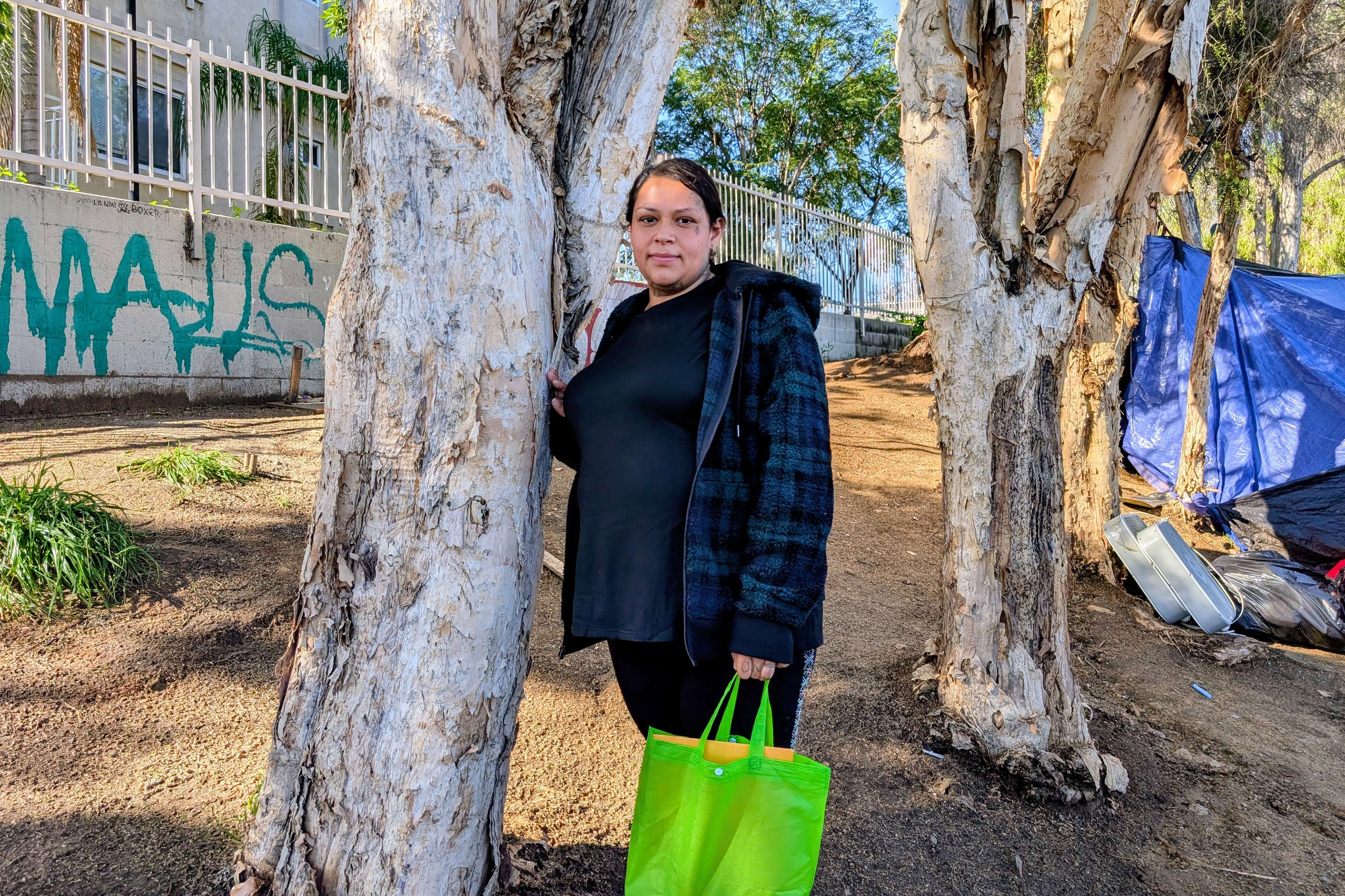 Mia Angulo stands by a tree holding a bright green bag. A homeless encampment is seen in the background behind her.