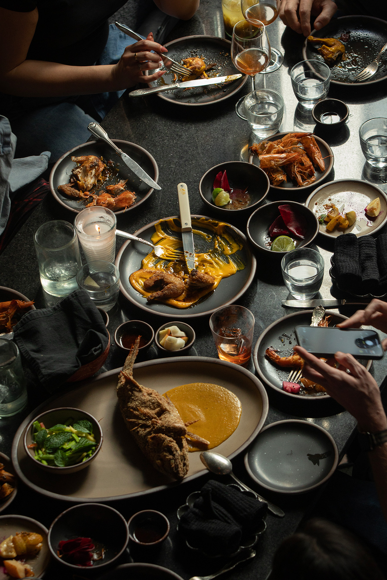 A spread of dishes on a black tabletop — included is a fried whole fish, head-on shrimp, and salad.