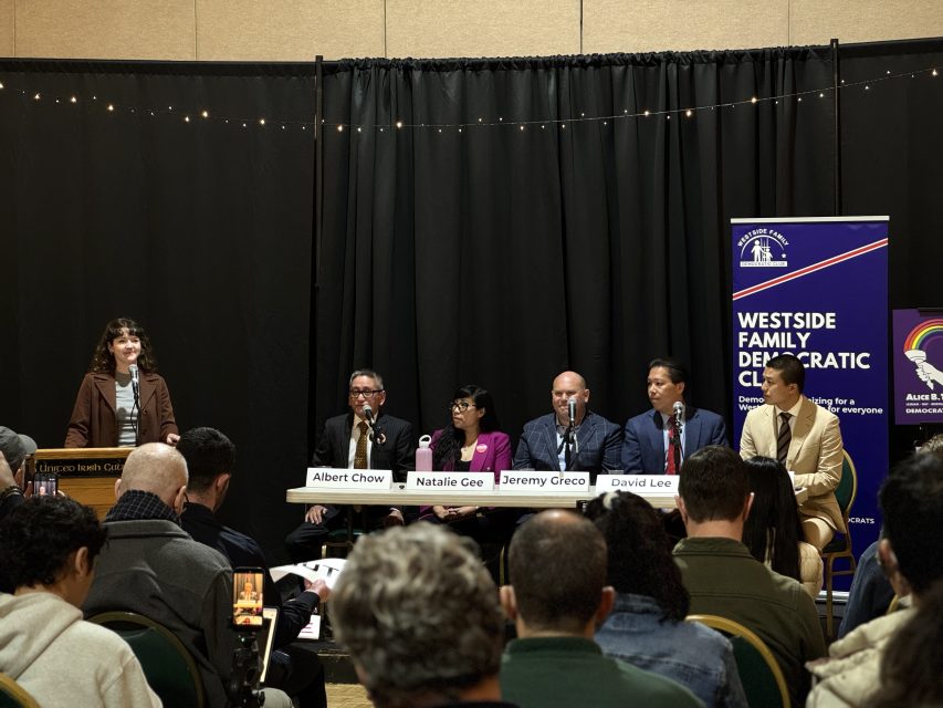 A panel of five speakers sits at a table on stage with name cards, while a woman stands at a podium addressing an audience at an indoor event.