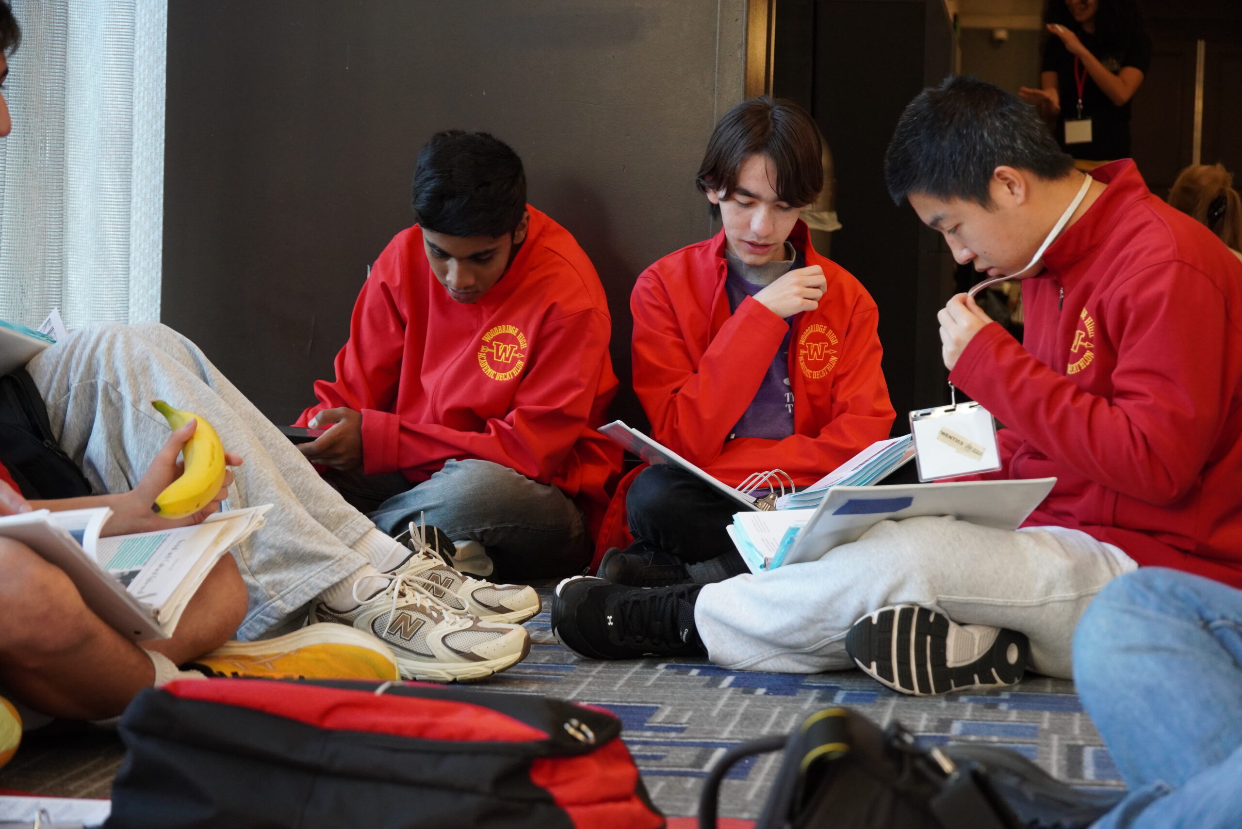 Students in red team jackets sit in a hallway studying notes and materials together during the Academic Decathlon competition.