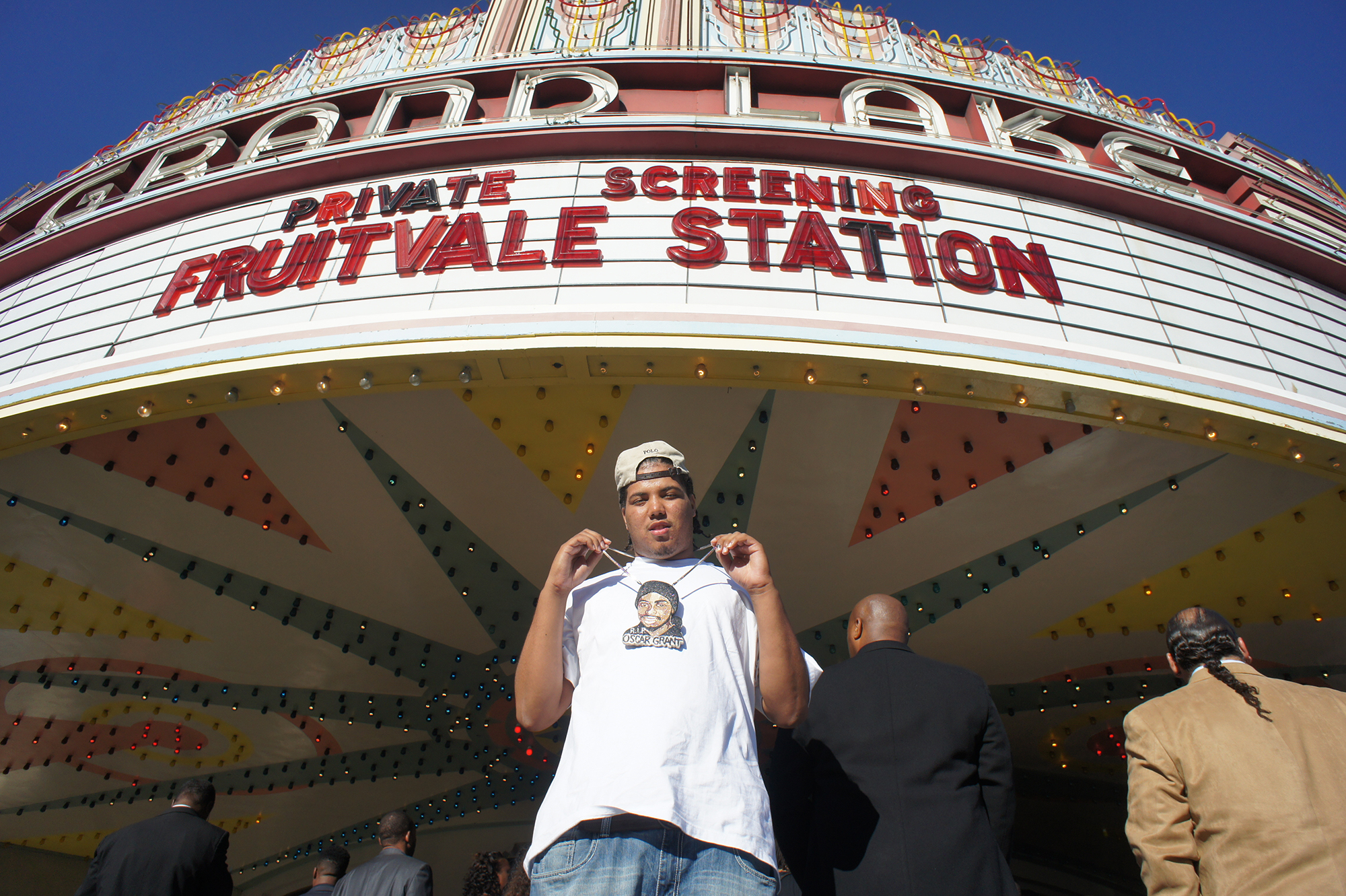 A man in a white cap and T-shirt holds up a medallion in the image of Oscar Grant, beneath a theater marquee reading 
