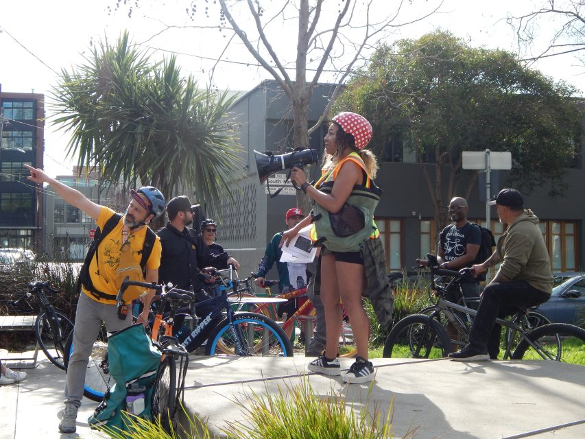 A group of cyclists gathers outdoors while a woman in a reflective vest and red helmet stands on a ledge with a megaphone; one man points to the left.