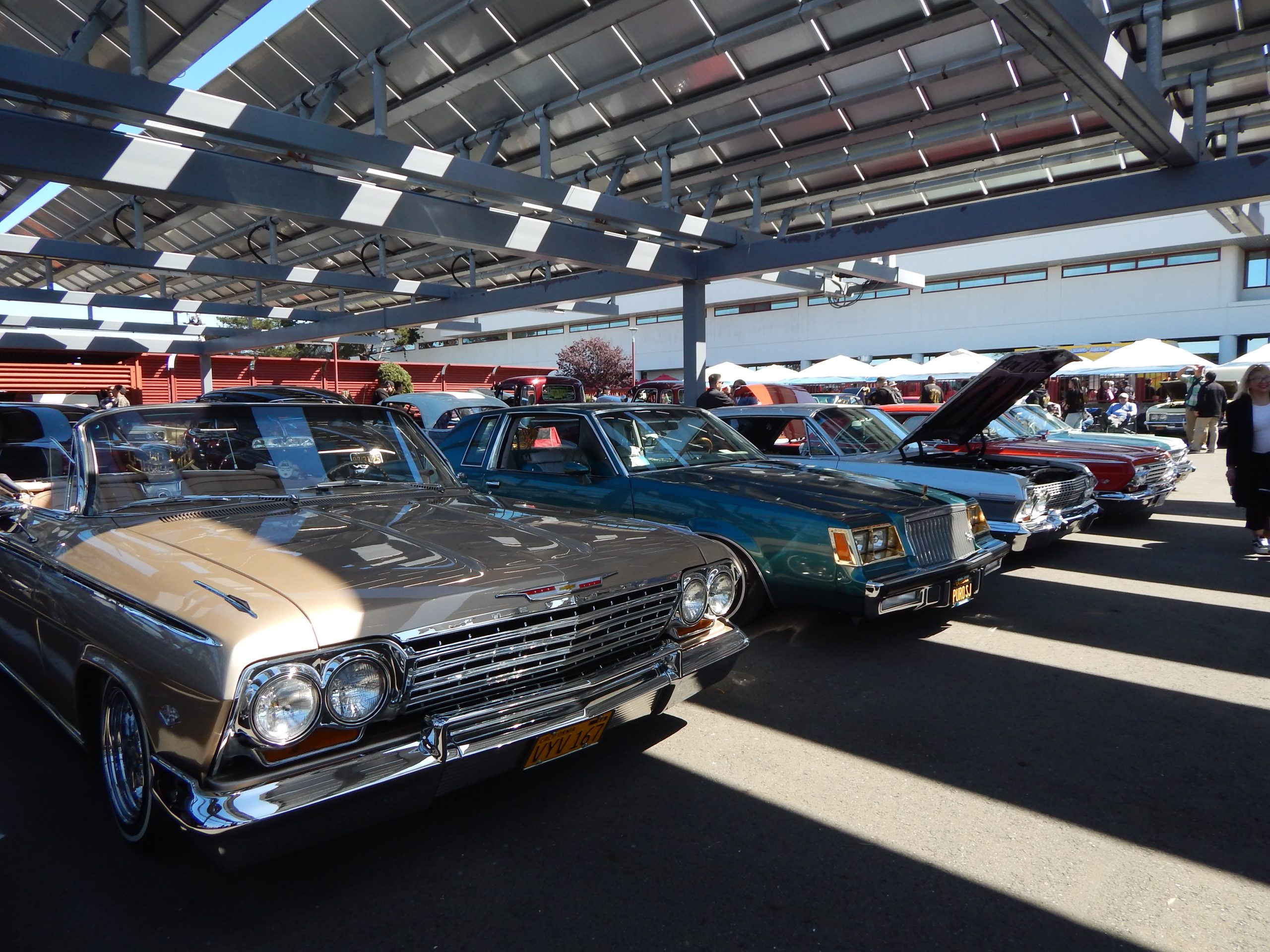 Several classic cars are parked in a row under a metal canopy at a car show, with people and tents visible in the background.