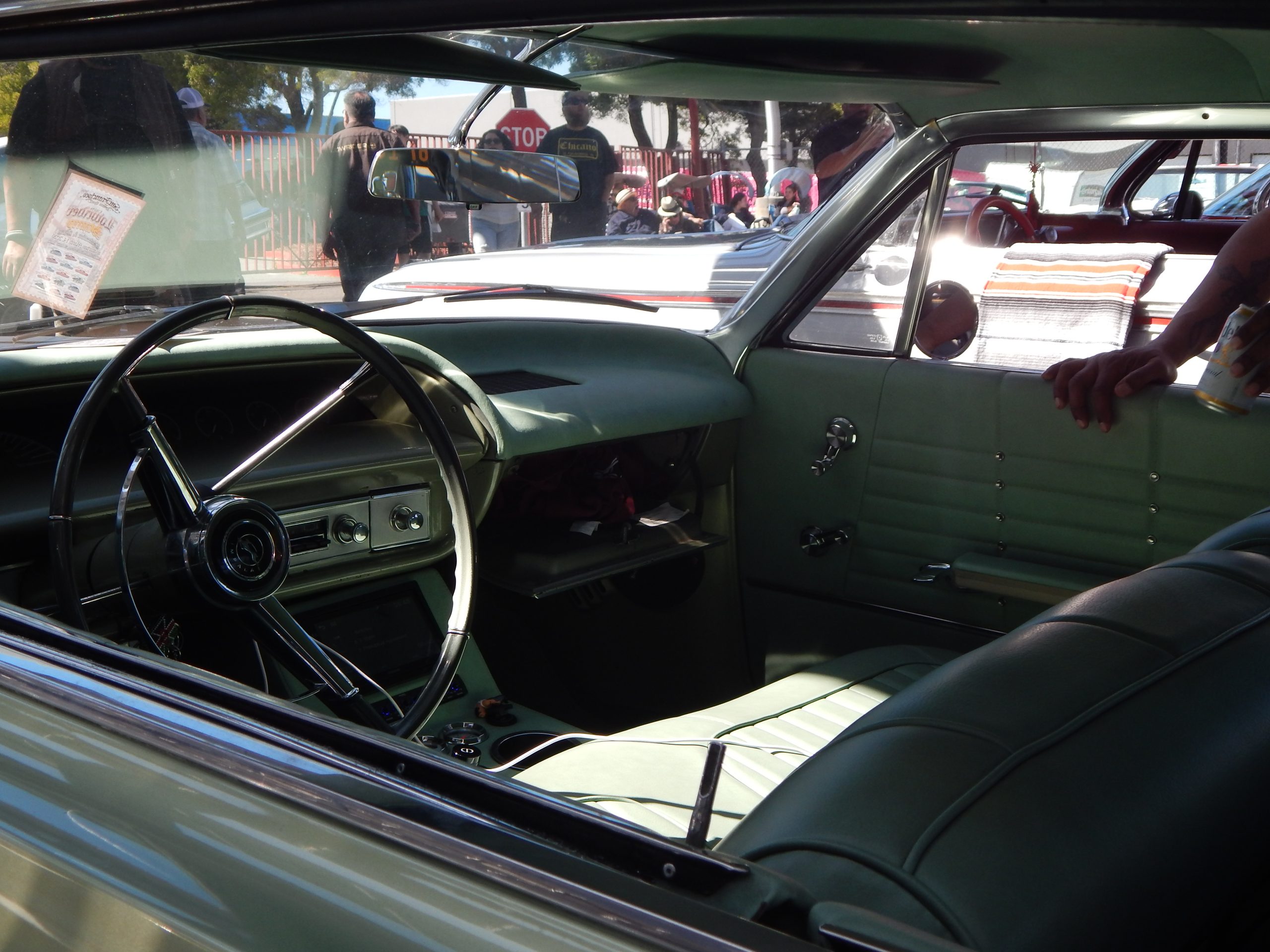 Interior view of a vintage car with green upholstery, showing the steering wheel, dashboard, and front seat, with people and other cars visible outside through the windows.