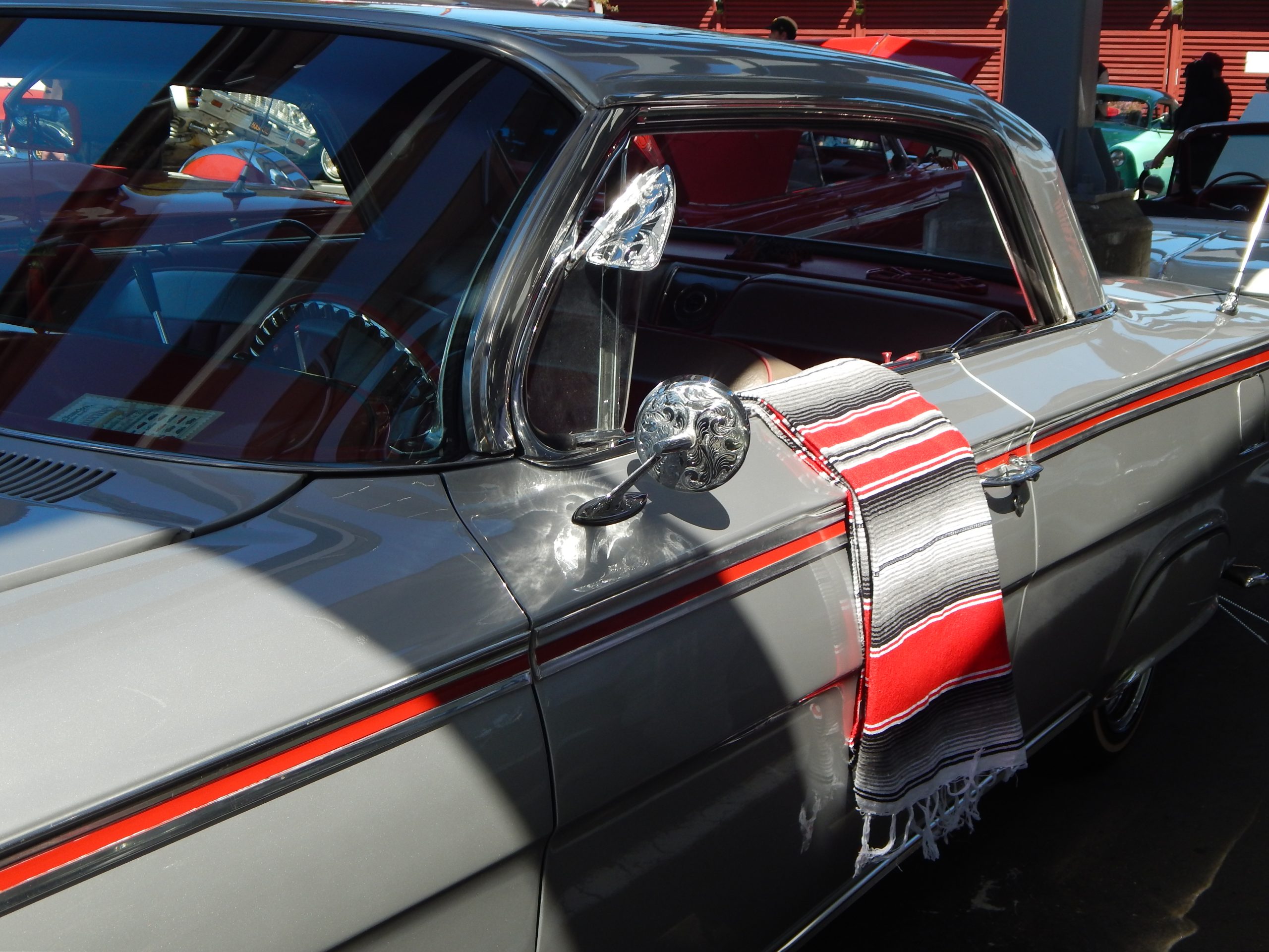 A gray vintage car with red and black accents displays a striped Mexican blanket draped over the passenger door at an outdoor car show.