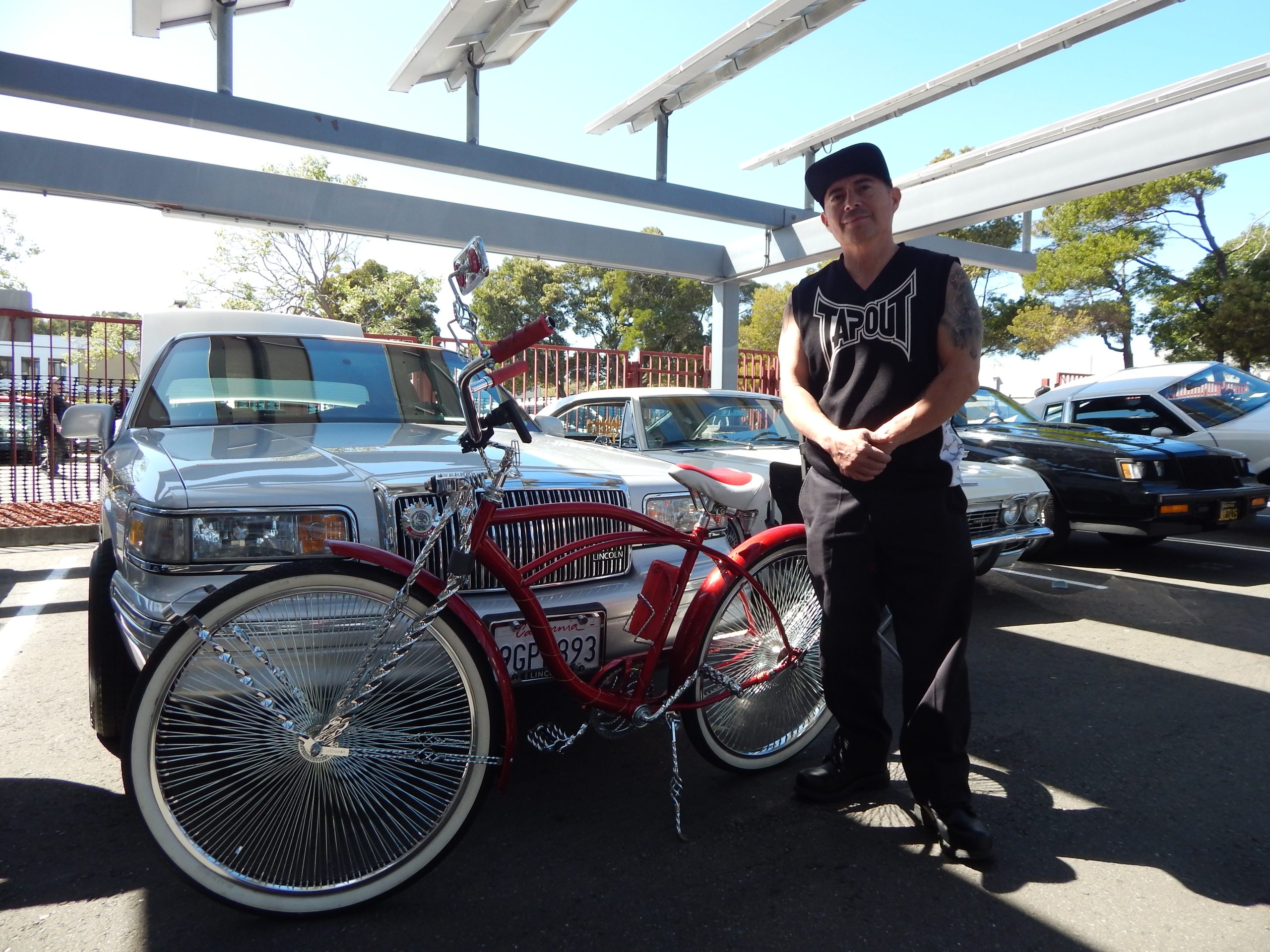 A man stands next to a red lowrider bicycle parked in front of a classic car in a parking lot under a metal canopy.