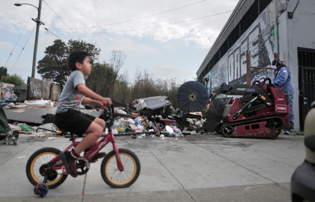 A young boy rides his bike along the sidewalk where Keep Oakland Beautiful and Clean workers, a division of Oakland Public Works department, removes a large trash pile along International Blvd. at 71st Avenue in Oakland, Calif., on Friday, Sept. 19, 2025. (Laura A. Oda/ Bay Area News Group)