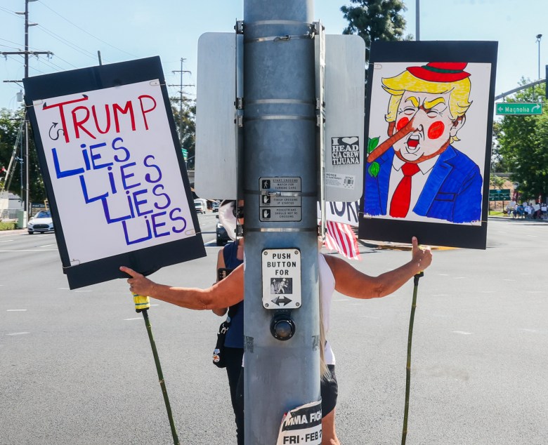 A protestor stands on a street corner holding two signs. One reads "Trump Lies" repeatedly; the other depicts a cartoon character with a long nose.