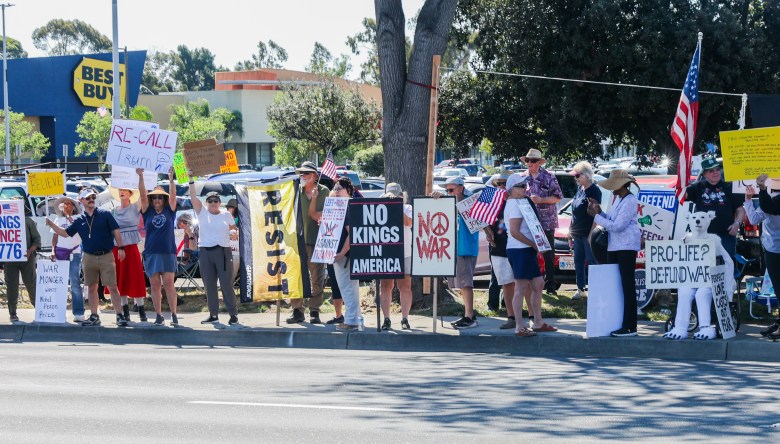 A diverse group of protesters stands on a sidewalk holding signs with messages like "No Kings in America" and "Pro-Life," conveying a peaceful demonstration.