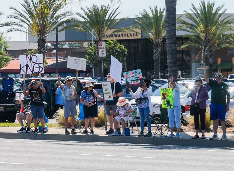 A group of protesters stands on a sidewalk holding signs with messages like "No Kings in America" and "No War." American flags are visible, set against a backdrop of parked cars.