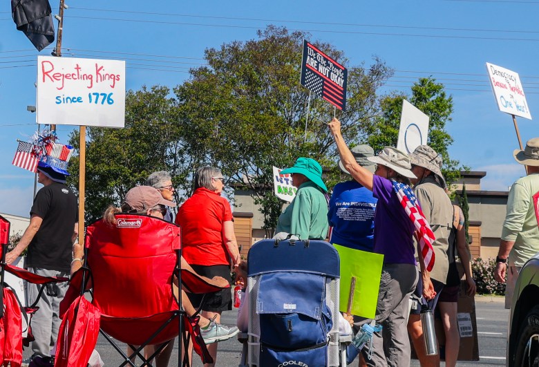 Protesters gathered outdoors, holding signs with phrases like "Rejecting Kings since 1776."