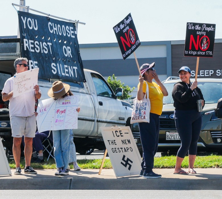 A group of protesters at a rally hold signs with messages against authority and ICE, invoking historical references. The mood is serious and determined.