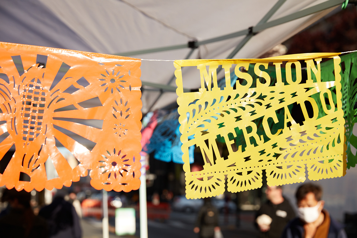 Colorful papel picado banners hang under a tent at an outdoor market. The yellow banner reads "Mission Mercado." People and city street are visible in the background.