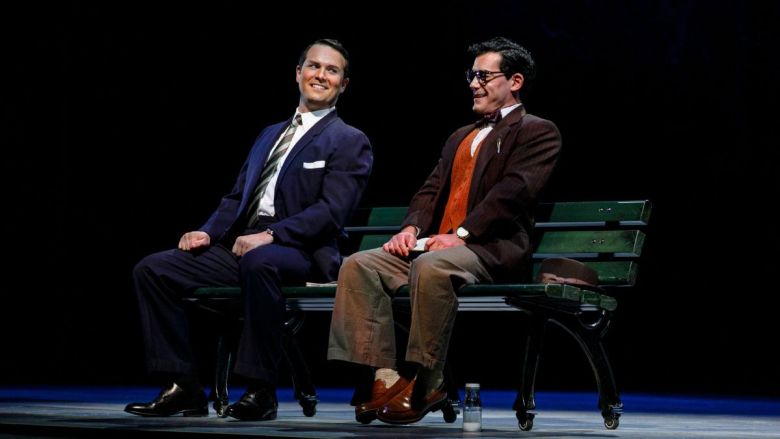 Two men in suits sit on a bench on stage, with a hat and bottle on the floor.