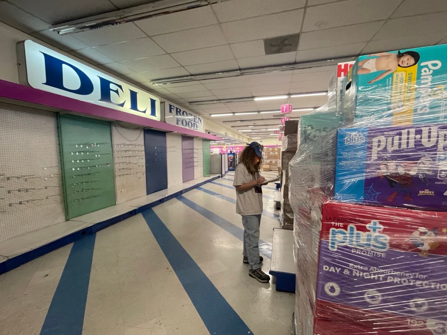 A person wearing a mask, a baseball hat, white t-shirt and jeans holds a clipboard in front of stacks of packages of diapers, with signs that read "Deli" and "frozen food" in the background.