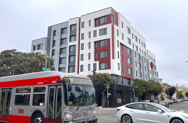 A red and gray bus and a white car pass in front of a multistory apartment building on the corner of 6th Ave and Geary Boulevard in San Francisco.
