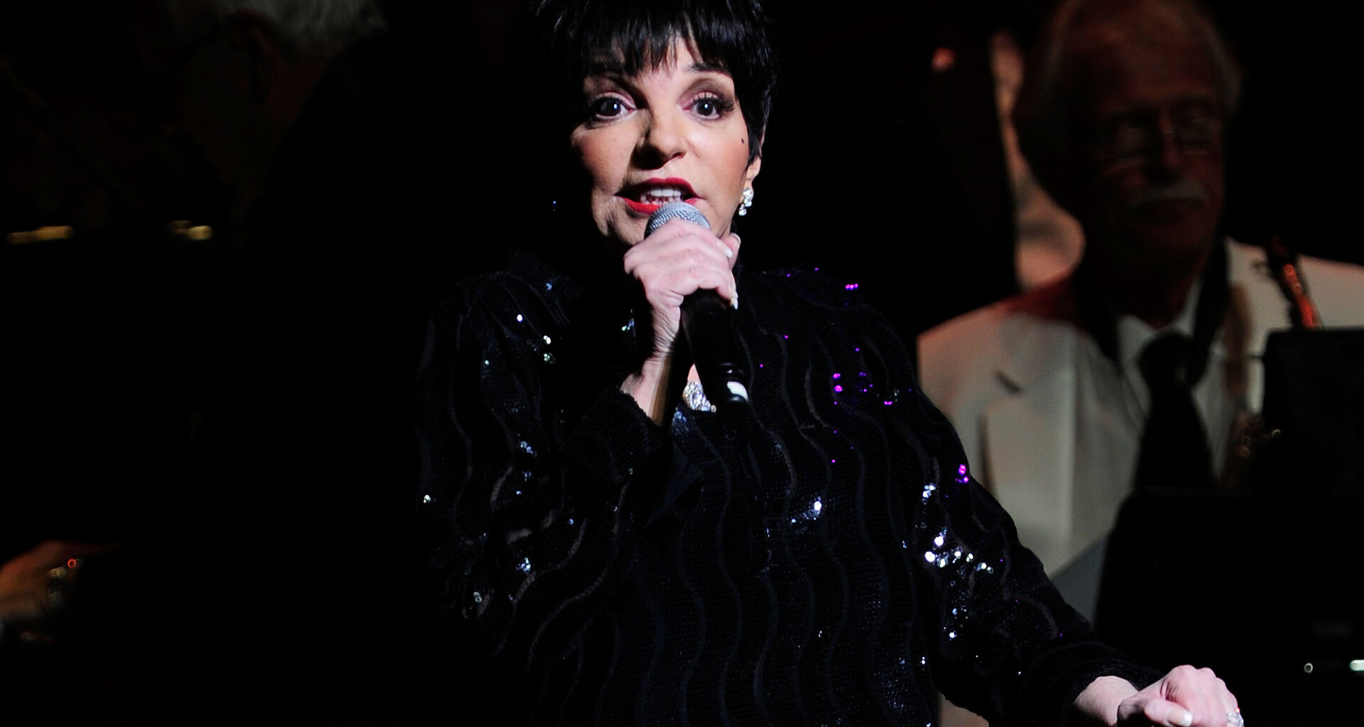 A woman with a black bob haircut and dressed in black sings into a microphone, looking slightly upward