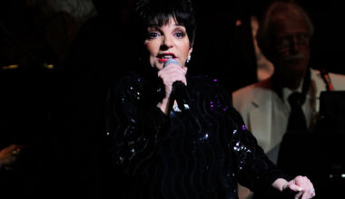 A woman with a black bob haircut and dressed in black sings into a microphone, looking slightly upward