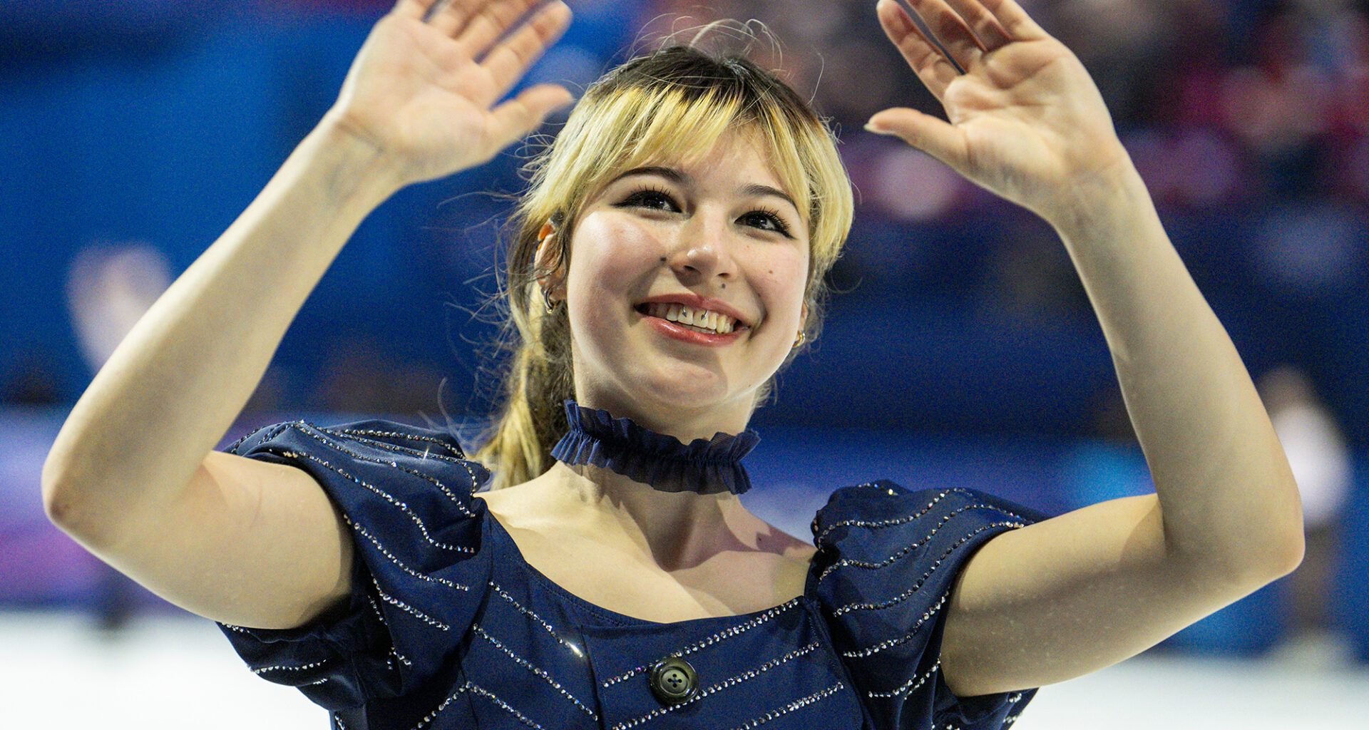 A young woman in a blue outfit and choker necklace waves to a crowd.