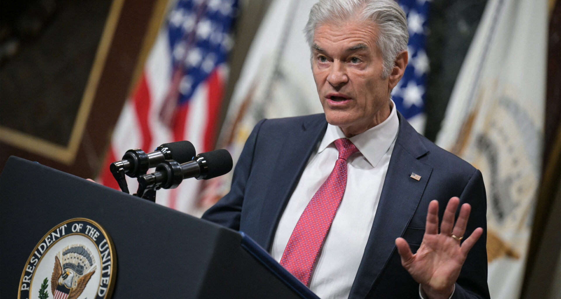 A photo of Mehmet Oz speaking at a podium at the White House. American flags are seen behind him.