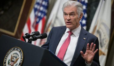 A photo of Mehmet Oz speaking at a podium at the White House. American flags are seen behind him.