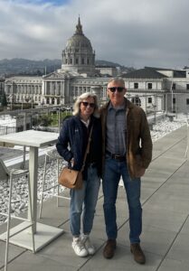 Jennifer and Bob Hagle, wearing sunglasses and warm clothes, pose in front of the Sky Deck of 333 Golden Gate Ave. with the San Francisco City Hall dome and building behind them.