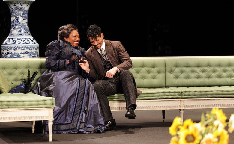 Two people laughing on a green sofa near a large blue and white vase.