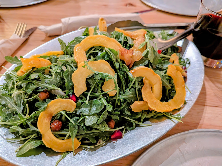 A salad with arugula, roasted squash slices, pomegranate seeds, and chickpeas is served on a metal platter next to a glass of dark beverage.