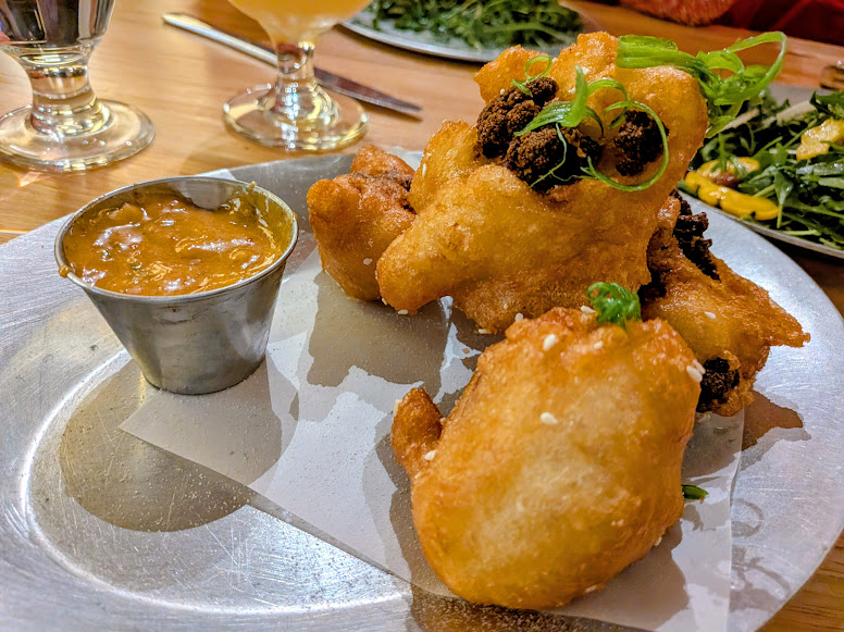 Three pieces of fried batter, garnished with green onions, are served on a plate with a small cup of dipping sauce and a side of greens in the background.