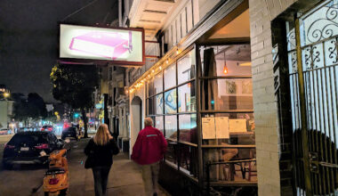 Two people walk on a city sidewalk at night past a lit restaurant with large windows and a pink sign above the entrance. A parked scooter is visible.