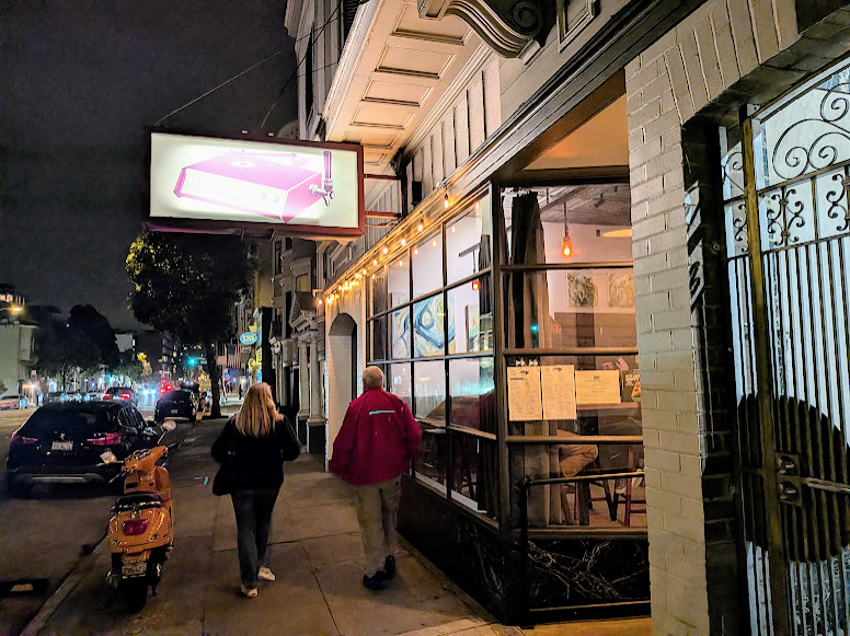 Two people walk on a city sidewalk at night past a lit restaurant with large windows and a pink sign above the entrance. A parked scooter is visible.