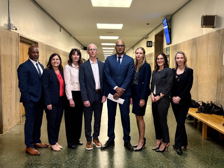 Eight professionally dressed individuals stand together in a hallway with beige walls and overhead lighting; a bench and bags are visible on the right.