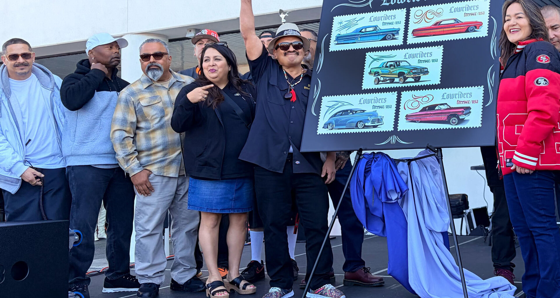A group of Latino celebrants gather around an easel showing five stamp designs of lowrider-style cars