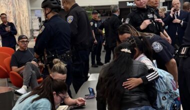 Police officers and several people, some sitting and some standing, gather in a public indoor area with cell phones and backpacks visible.
