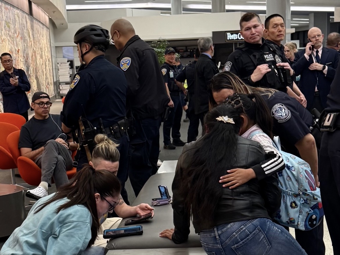 Police officers and several people, some sitting and some standing, gather in a public indoor area with cell phones and backpacks visible.