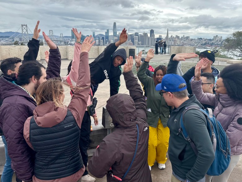 people gather in a circle on a patio raising their hands, with the San Francisco skyline in the background