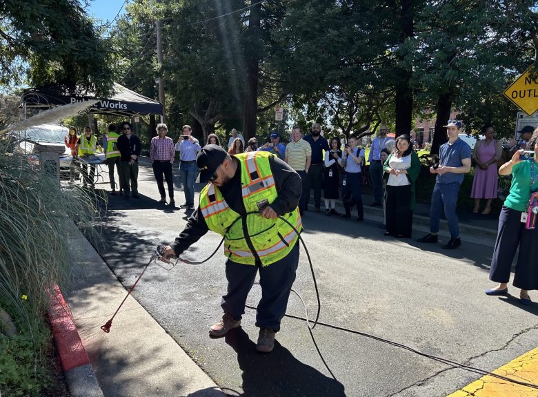 A worker in a yellow safety vest sprays red paint onto a curb while a crowd of people looks on, many of them taking photos on cellphones